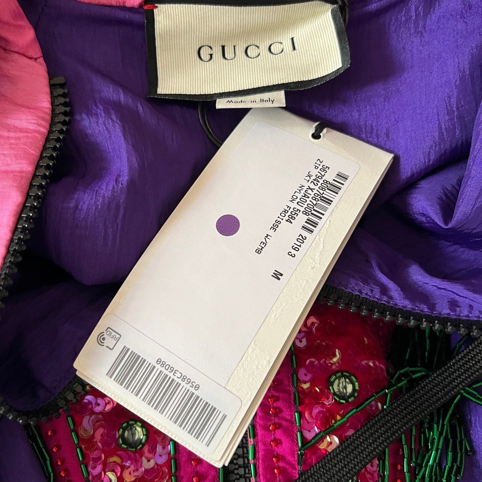 Close-up of a purple Gucci garment interior with a visible Gucci neck label and a product tag, showing pink and magenta sequined fabric with black zipper trim.
