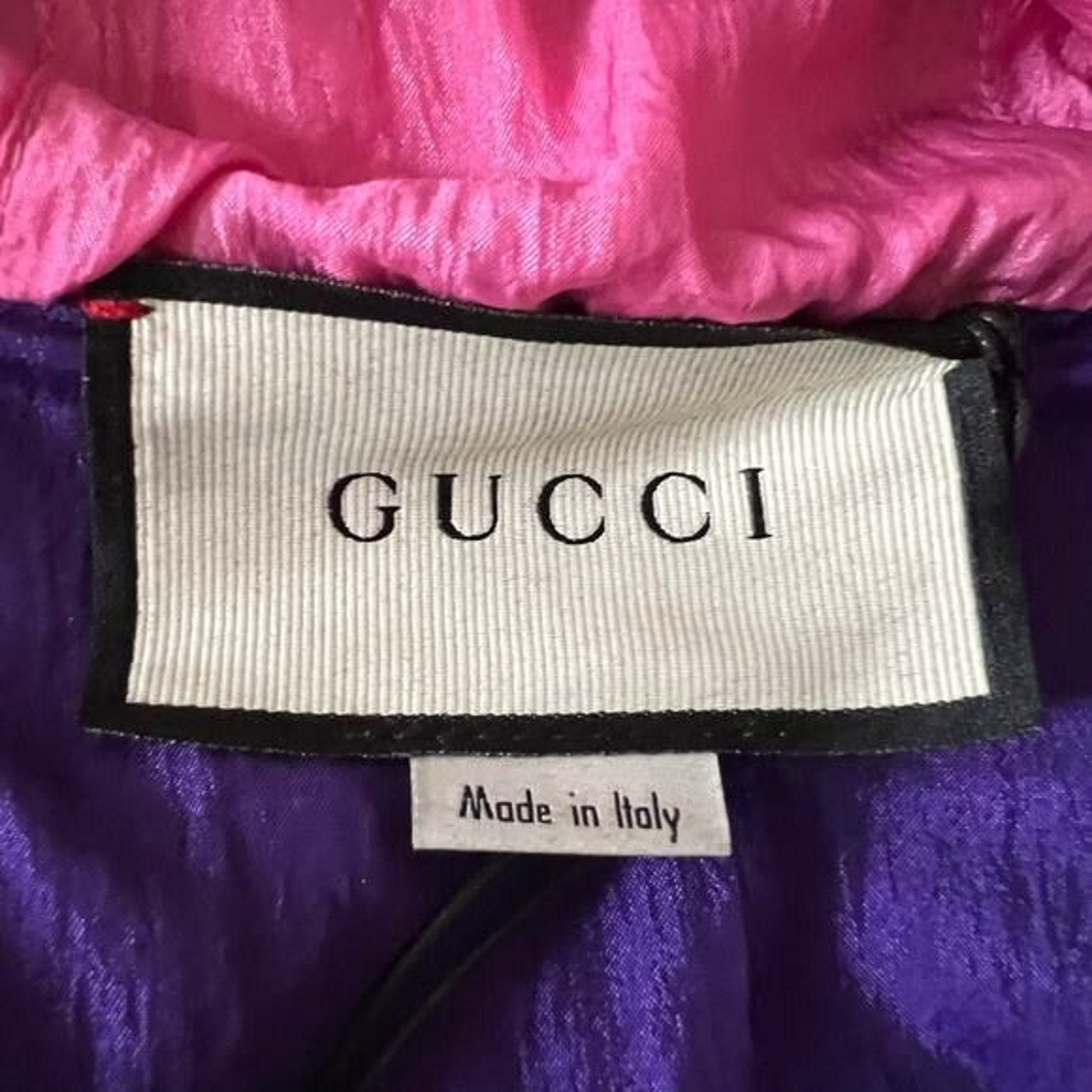 Close-up of a Gucci garment label on a purple and pink fabric item, with the Gucci brand name clearly visible on the white label.
