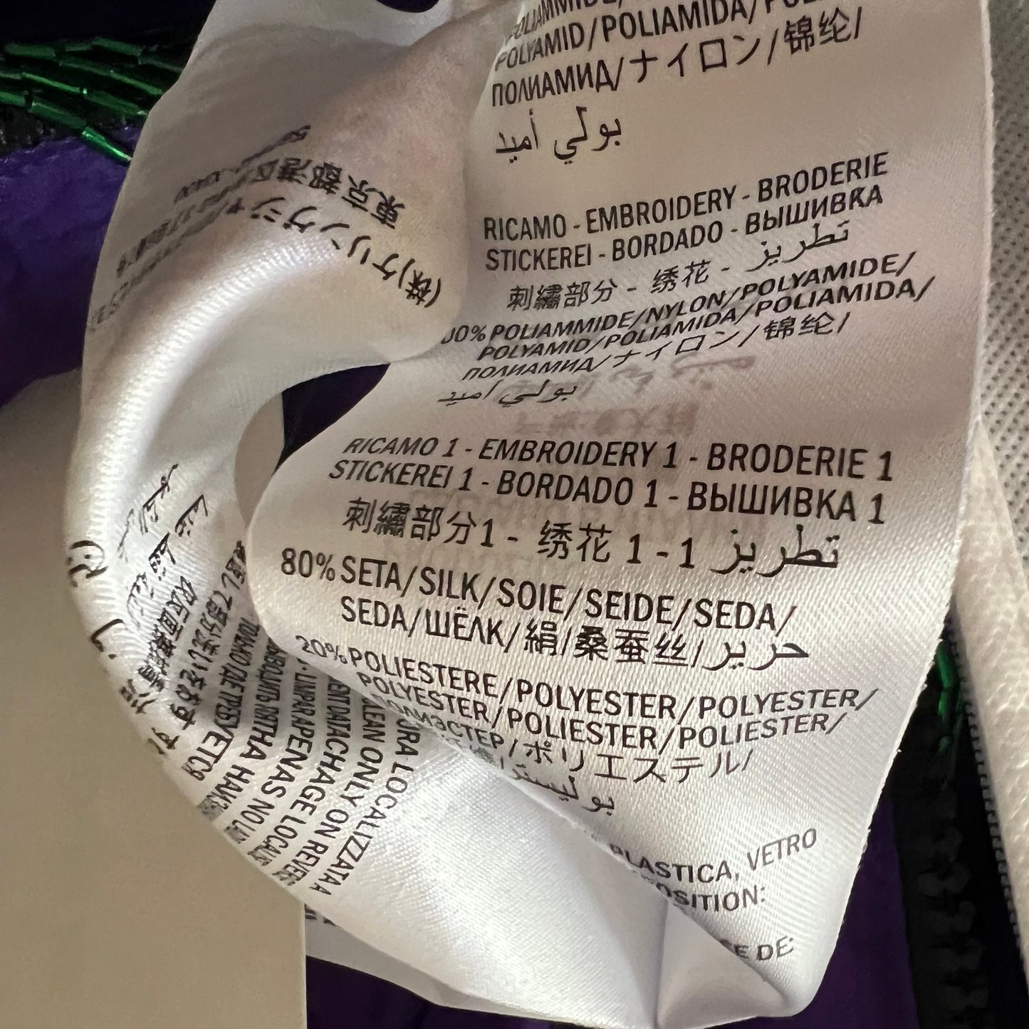 Close-up of a white fabric care label with printed text including 'RICAMO - EMBROIDERY - BRODERIE' and material information, photographed against a dark green and purple textured background.