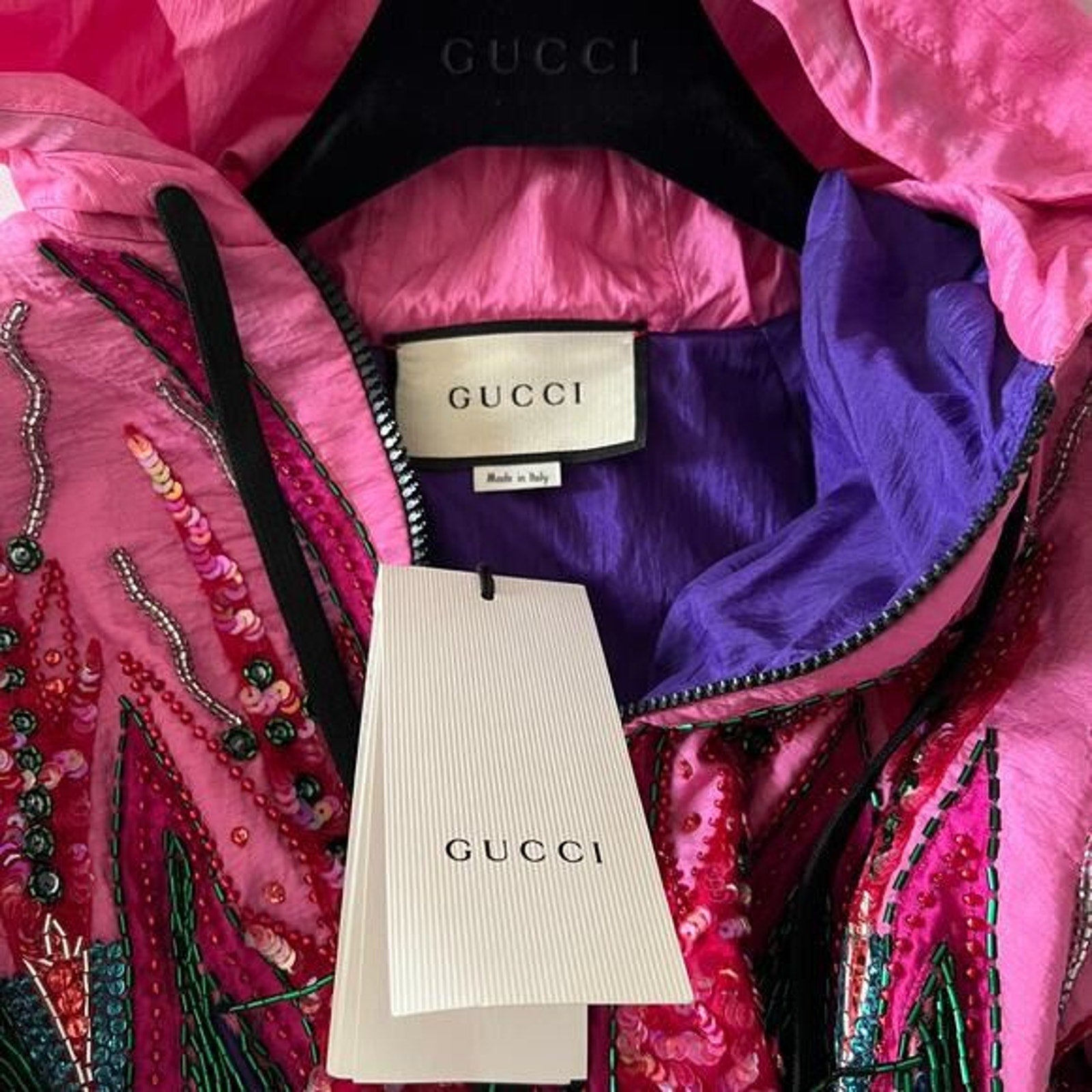 A close-up of a Gucci pink embroidered jacket or windbreaker on a black hanger, showing a purple inner lining, a cream Gucci neck label, and a hanging Gucci price tag.